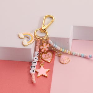May include: A gold-colored keychain with a rainbow, flower, heart, star, and seashell charms. The heart charm says "I'M GOOD". The keychain also has the name "RILEY" spelled out with white letter beads. The background is pink and white.