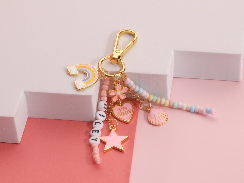 May include: A gold-colored keychain with a rainbow, flower, heart, star, and seashell charms. The heart charm says "I'M GOOD". The keychain also has the name "RILEY" spelled out with white letter beads. The background is pink and white.