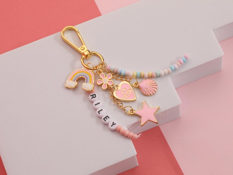 May include: A gold-colored keychain with a rainbow, flower, heart, seashell, and star charms. The heart charm says "It's Good". The keychain also has a beaded strand and a strand of beads spelling "RILEY".