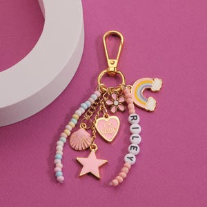 May include: A gold-tone keychain with multiple charms, including a pink heart, star, and seashell. The keychain also features a rainbow charm and a beaded strand spelling "RILEY". The background is a vibrant pink.