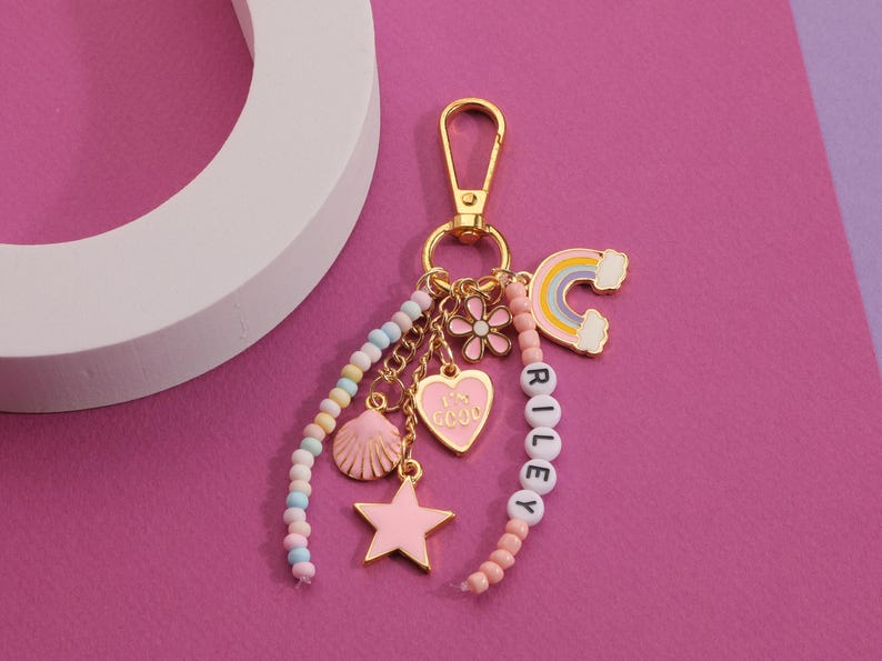 May include: A gold-tone keychain with multiple charms, including a pink heart, star, and seashell. The keychain also features a rainbow charm and a beaded strand spelling "RILEY". The background is a vibrant pink.