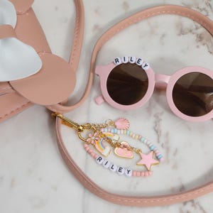May include: A pink purse with a white bow, round pink sunglasses with the name "RILEY" on the frame, and a beaded bracelet with charms. The sunglasses have a white daisy on the side. The bracelet also has the name "RILEY".