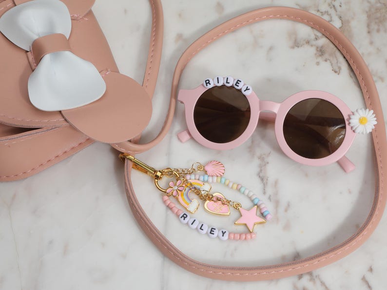 May include: A pink purse with a white bow, round pink sunglasses with the name "RILEY" on the frame, and a beaded bracelet with charms. The sunglasses have a white daisy on the side. The bracelet also has the name "RILEY".