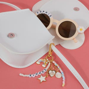 May include: A white purse with a matching strap, next to a pair of light pink sunglasses with the name "SOPHIE" spelled out on the frame. A gold keychain with a rainbow, star, and heart charm is attached.