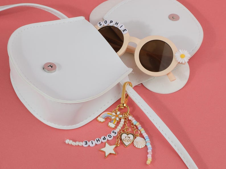 May include: A white purse with a matching strap, next to a pair of light pink sunglasses with the name "SOPHIE" spelled out on the frame. A gold keychain with a rainbow, star, and heart charm is attached.