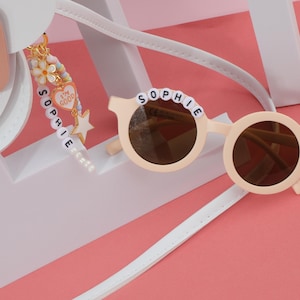 May include: Round, light beige sunglasses with brown lenses, the name "SOPHIE" spelled out in white beads across the top. A daisy decorates one side. A charm with the words "I'm good" hangs from a white strap. The background is pink and white.
