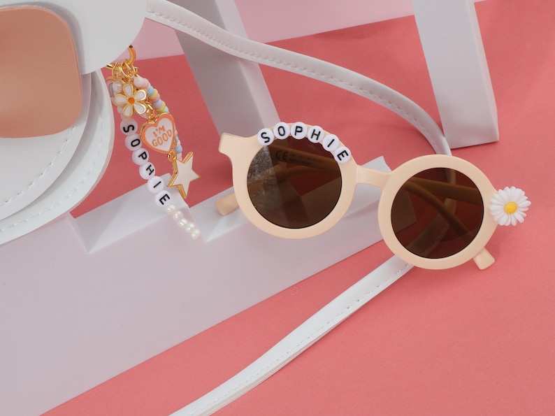 May include: Round, light beige sunglasses with brown lenses, the name "SOPHIE" spelled out in white beads across the top. A daisy decorates one side. A charm with the words "I'm good" hangs from a white strap. The background is pink and white.