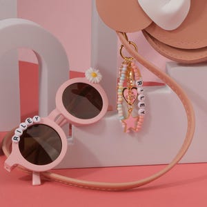 May include: Pink round sunglasses with the name "RILEY" spelled out in white letter beads. A daisy decorates the top of one lens. A charm bracelet with the name "RILEY" and a pink star charm hangs from a pink purse.
