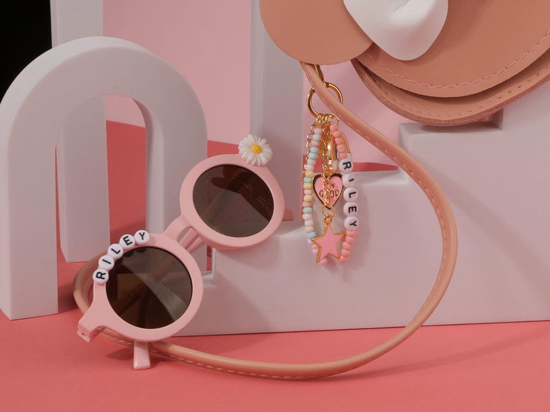 May include: Pink round sunglasses with the name "RILEY" spelled out in white letter beads. A daisy decorates the top of one lens. A charm bracelet with the name "RILEY" and a pink star charm hangs from a pink purse.