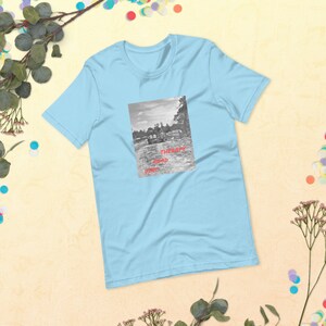 May include: Light blue t-shirt featuring a black and white photograph of a car driving down a road. The text "Therapy is Road Trip" is printed in red on the shirt.