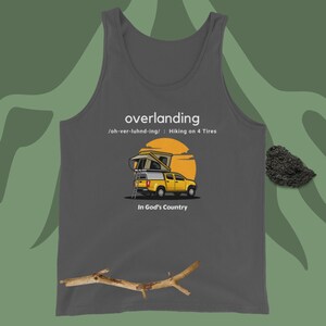 May include: A grey tank top featuring a graphic of a yellow pickup truck with a roof top tent. The text "overlanding" is printed in white above the truck, and the text "/oh-ver-luhnd-ing/ : Hiking on 4 Tires" is printed in white below the truck. The text "In God's Country" is printed in white below the truck.