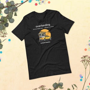 May include: A black t-shirt with a white graphic of a yellow truck with a pop-up camper. The text "overlanding" is written in white above the truck, with the definition "(ah-ver-luhnd-ing): Hiking on 4 Tires" below the truck. The text "In God's Country" is written in white below the truck.