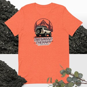May include: Orange t-shirt featuring a graphic of a white off-road vehicle driving through a mountain landscape. The text "Dirtroad Therapy" and "In God's Country" is printed below the vehicle.