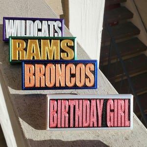 May include: Four colorful rectangular signs with different team names. The signs are arranged vertically, with the top sign reading "WILDCATS", the second sign reading "RAMS", the third sign reading "BRONCOS", and the bottom sign reading "BIRTHDAY GIRL".