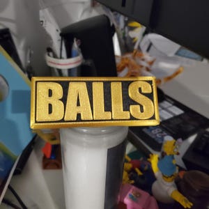 May include: A gold-colored sign with the word "BALLS" in black letters. The sign is made of a textured material and has a slightly raised edge.
