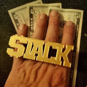 May include: A gold-colored ring spelling out "STACK" is held in front of several five-dollar bills. The ring is the focal point, with the word's letters prominently displayed. The background is a dark, textured surface.
