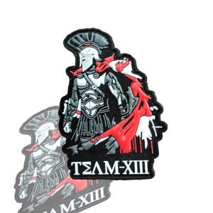 May include: A black patch featuring a Spartan warrior in a helmet and armor, with a red cape. The text "TEAM-XIII" is displayed in white. The patch has a detailed design and a shadow effect.