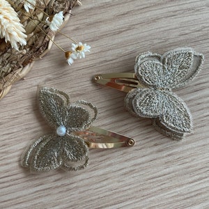 Peut inclure: Deux pinces à cheveux dorées avec des motifs de papillons brodés en fil doré. L'une des pinces est ornée d'une petite perle. Elles sont posées sur une surface en bois clair.