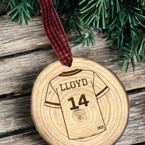 May include: A wooden Christmas ornament with a football jersey design. The jersey is engraved with the name "LLOYD" and the number "14". A red and black plaid ribbon is attached for hanging. The ornament is set against a backdrop of pine needles and wood.