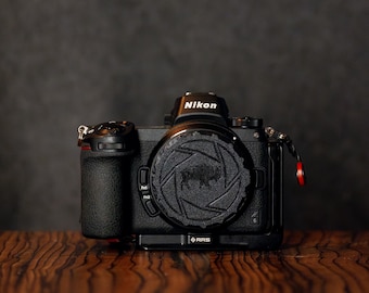 May include: A black Nikon camera with a black lens cap featuring a buffalo silhouette. The camera is sitting on a wooden surface.