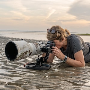 May include: A person lies on the sand, using a camera with a long lens to take a photo. The camera is mounted on a tripod and the lens is pointed towards the horizon. The sand is wet and there are small puddles of water on the surface.