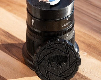 May include: A black camera lens with a black lens cap featuring a buffalo silhouette. The lens is labeled "Nikon 14-30/4 S".