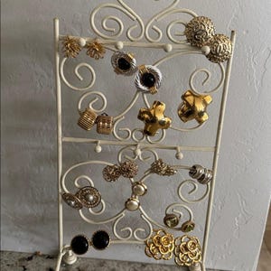 May include: An off-white metal earring display stand holding a variety of vintage earrings. The earrings are gold, silver, and black, featuring various designs like floral, geometric, and pearl accents. The stand has a scrolled design.