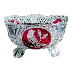 May include: A clear cut glass bowl with a scalloped rim and three feet. The exterior features a diamond pattern and a central oval medallion in deep red glass, displaying an etched design of a bird perched next to a sprig of wheat.