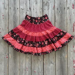 May include: A tiered skirt featuring a repeating floral pattern in shades of black, deep red, and bright pink. The garment is constructed with multiple horizontal fabric bands, each showcasing a different variation of the floral print and color scheme.