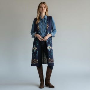 May include: A long, navy blue vest with a Southwestern-inspired geometric pattern in shades of teal, cream, and brown. The vest is worn over a denim shirt and dark jeans, paired with brown cowboy boots. The vest's design features repeating geometric shapes.
