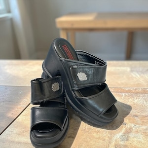 Peut inclure: Sandales compensées en cuir noir avec des brides réglables. Les sandales arborent le logo Harley-Davidson sur les brides et la semelle intérieure. Le design à bout ouvert et la semelle épaisse offrent un look audacieux.