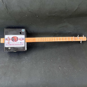 May include: A black cigar box guitar with a Donald J. Trump "Limited Edition" sticker on the front. The guitar has a wooden neck and six tuning pegs.