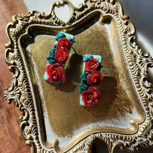 May include: A pair of red rose earrings with green leaves on a turquoise background. The earrings are displayed on a gold tray.
