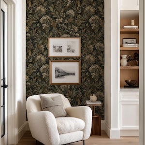 May include: A cream-colored bouclé armchair with a striped throw pillow sits in front of a wall covered in dark botanical wallpaper featuring white and green floral patterns. Two framed landscape prints hang above the chair.