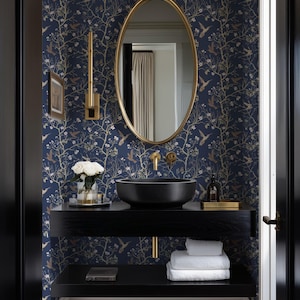 May include: A modern bathroom with a navy blue wallpaper featuring a bird and floral pattern. A black sink sits atop a black floating vanity with gold fixtures. A gold-framed oval mirror hangs above the sink, reflecting the room. White towels are stacked on a lower shelf.