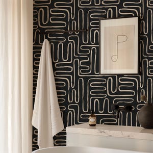 May include: A black wallpaper featuring a continuous white hand-drawn maze-like pattern. A white towel hangs on a black bar, a framed line art print hangs on the wall, and a small brown bottle sits on a white marble vanity next to a black vase.