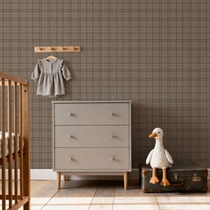May include: A nursery scene featuring a gray dresser with three drawers, a wooden crib, and a plush white goose toy. A light gray dress hangs on a wooden peg rack against a plaid wallpaper background. The room has a warm, neutral color palette.