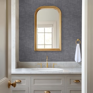 May include: A bathroom vanity with a gold-framed arched mirror, gold faucet, and hardware. The vanity has gray cabinets and a white countertop. A white towel hangs from a gold towel ring. The wall is covered in a textured gray wallpaper.