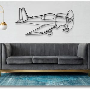 May include: A black metal wall sculpture of a small airplane with a propeller. The sculpture is mounted on a white wall above a gray sofa with gold legs. The sofa is in front of a blue rug.