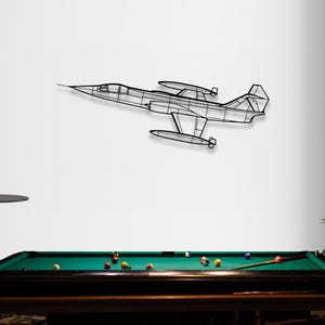 以下が含まれることがあります： 白い壁に黒い金属製のジェット機のワイヤーフレームが取り付けられています。飛行機は、翼、胴体、尾を示す詳細なアウトラインです。下には、ビリヤードボールとキューを備えた緑色のビリヤード台が見えます。
