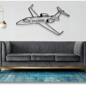 May include: A black metal wall sculpture of a private jet, with a gray sofa in the foreground. The sofa has a gold base and is in front of a white wall.