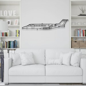 May include: A black metal airplane wall art piece. The airplane is a side profile of a jet, with detailed windows and wings. It is mounted on a white wall above a white couch.