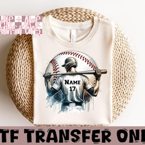 May include: A white t-shirt with a baseball player graphic. The player is wearing a baseball uniform with the number 17 and the word "NAME" on the front. The player is holding a baseball bat and is standing in front of a baseball.