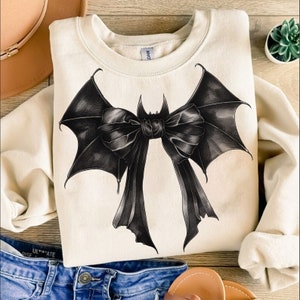 May include: Cream-colored sweatshirt featuring a detailed black bat bow design. The bow has large, textured wings and flowing ribbons. The sweatshirt is folded, with a glimpse of denim jeans and a hat in the background, suggesting a casual style.