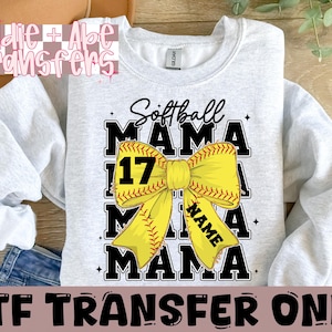 May include: A gray sweatshirt with a softball design. The design features a yellow bow with a softball in the center and the text "Softball Mama" above and below the bow. The number "17" is on the softball. The text "NAME" is in the center of the bow.