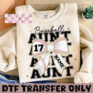 May include: A beige sweatshirt with a black and white design that says "Baseball Aunt" with a white bow and a baseball with the number 17 on it. The design also includes the text "NAME" in a gold font.