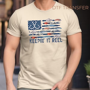 May include: Beige t-shirt featuring a graphic design of an American flag made of fishing lures and hooks. The text "KEEPING IT REEL" is printed in blue below the flag design. The shirt is a casual wear item.