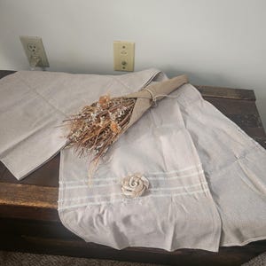 May include: A beige linen table runner with a decorative white trim and a fabric flower. A bundle of dried flowers wrapped in brown paper rests on the runner. The runner is displayed on a dark wooden surface.