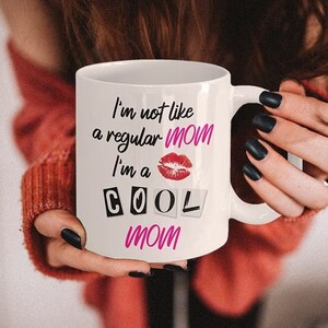 A stylish mom wearing a sweater with long dark hair and black nail polish, holding the "I'm Not Like a Regular Mom, I'm a Cool Mom" mug. Her relaxed style captures the fun and lighthearted spirit of this coffee cup, making it a perfect gift for cool moms everywhere.