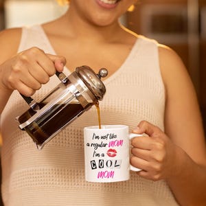 A stylish mom wearing cozy loungewear holds the "I'm Not Like a Regular Mom, I'm a Cool Mom" mug while pouring coffee. Her joyful expression captures the fun and lighthearted spirit of this coffee cup, making it a perfect gift for cool moms everywhere.